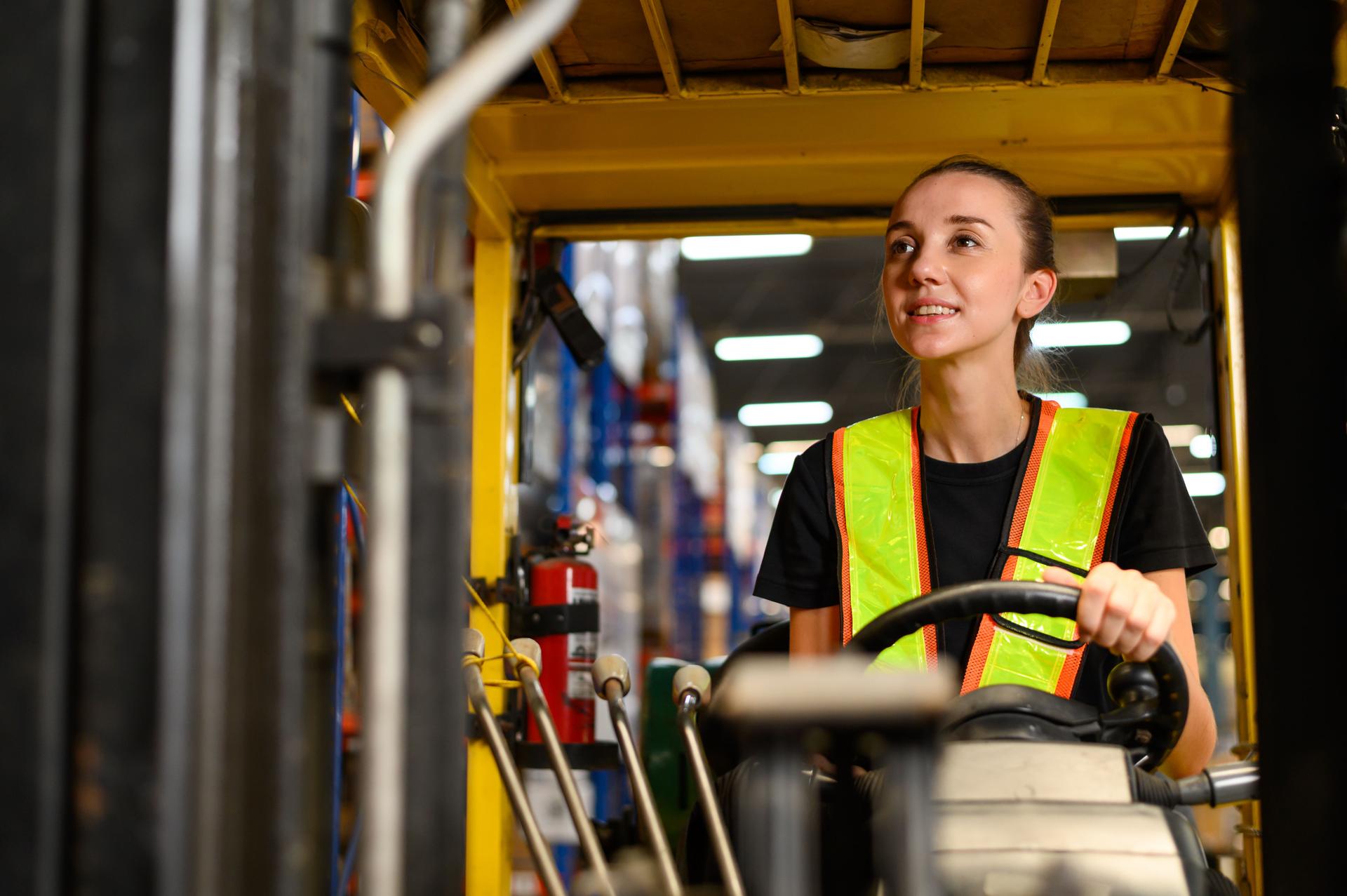 Young female warehouse worker driving a forklift at shipping warehouse. Industrial engineer woman in safety vest drives reach stacker to lift cargo box at freight cargo warehouse port.