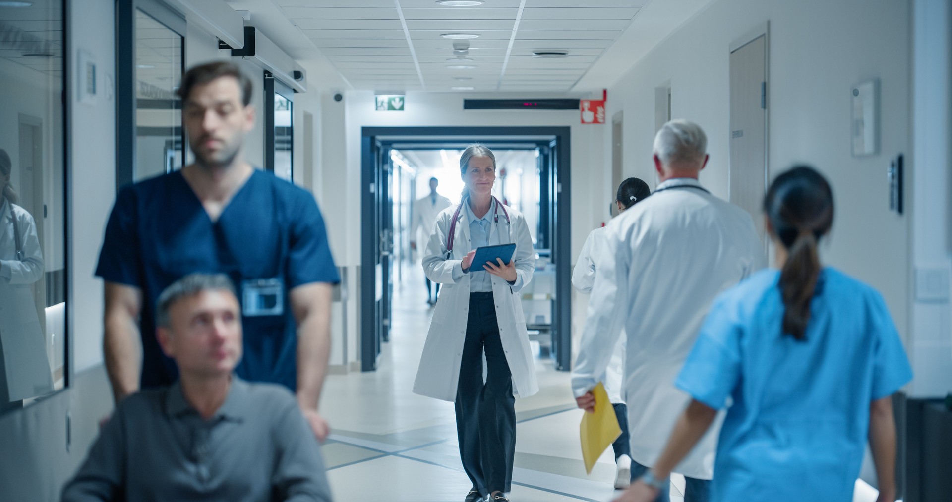 Diverse Medical Staff Walking Down a Hospital Hallway, Each Headed Towards Different Departments. Caucasian Female Doctor Discussing Treatment Plans, Reviewing Patient Charts on a Tablet